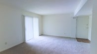This single-story space features a clean, minimalist design centered around an open living area. The room is finished with wall-to-wall neutral-toned carpeting, providing a soft and comfortable foundation throughout. Crisp white walls and a smooth white ceiling create a bright, airy atmosphere that feels open and welcoming.

A prominent sliding glass door, fitted with vertical blinds, serves as the main source of natural light, allowing sunlight to filter into the space and illuminate the room. The door likely leads to an outdoor area, offering potential for additional ventilation and outdoor access.

A wall-mounted HVAC vent is visible near the upper corner of the room, indicating a built-in climate control system for year-round comfort. The layout of the living area is straightforward and versatile, providing ample room for a variety of furniture arrangements. The neutral color palette of the walls, ceiling, and flooring creates a cohesive, understated aesthetic that serves as a blank canvas, easily adaptable to a range of interior design styles. Overall, the space presents a functional and well-lit environment with a simple, clean character.