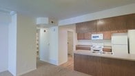 This single-floor space features an open-concept layout that connects the kitchen and living areas in a functional, easy-to-navigate design. The kitchen is equipped with warm wood-toned cabinetry spanning both upper and lower sections, providing generous storage. A built-in microwave is mounted above a white electric range, while a double sink with a standard faucet sits along the countertop peninsula that subtly divides the kitchen from the carpeted living area. The countertops offer a neutral tone that complements the cabinetry. The flooring transitions from hard surface in the kitchen and hallway areas to plush carpet in the main living space. A structural soffit above the kitchen adds architectural definition to the ceiling. Moving toward the rear of the space, a hallway leads to additional rooms, with a door opening to what appears to be a laundry or utility closet with shelving visible inside. A set of built-in open shelves lines one wall of the hallway, offering convenient storage or display space. Neutral wall tones and standard overhead HVAC vents contribute to a clean, practical interior throughout.