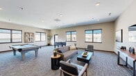Located at 2109 E Anamosa St in Rapid City, South Dakota, this generously sized single-floor community living space offers a well-appointed and versatile gathering area designed for recreation and relaxation. The open layout is anchored by plush, patterned carpeting and neutral-toned walls that create a clean, modern aesthetic throughout.

The living area is thoughtfully divided into distinct activity zones. A comfortable seating arrangement features a contemporary sofa, accent chairs, and coordinating coffee and side tables, providing an inviting space for socializing. A wall-mounted flat-screen TV with a console table and tasteful artwork adds a media lounge element to the room.

The game and recreation area is a standout feature, housing a full-size pool table and a foosball table, making it ideal for community entertainment. Recessed lighting throughout the ceiling ensures consistent, well-distributed illumination across all zones.

Multiple large, dark-framed windows line the walls, flooding the interior with abundant natural light and offering views of the surrounding South Dakota hillside landscape and outdoor fenced areas. A direct exterior access door provides convenient connectivity to the outdoors.

Decorative touches, including framed wall art and potted greenery, contribute warmth and character to the space. The overall design strikes a balance between functional recreation and comfortable community living.