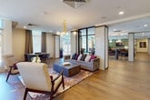 This expansive single-floor amenity lounge is designed with a sophisticated mid-century modern aesthetic, seamlessly blending functionality with refined style. Warm hardwood flooring flows throughout the entire space, unifying a series of well-appointed zones distinguished by thoughtful architectural dividers featuring textured white tile and dark metal-framed glass partitions.

The main lounge area anchors the space with plush velvet sofas, tufted seating, and mid-century accent chairs arranged around substantial wood coffee tables atop patterned area rugs. A sculptural gold-toned chandelier with globe pendants serves as a striking focal point overhead. Large windows and glass doors admit generous natural light and provide outdoor views throughout.

Adjacent to the lounge, a long bar counter lined with leather barstools is illuminated by elegant pendant lighting. A fully equipped kitchen features a generous white quartz island, dark countertops, wood cabinetry, stainless steel appliances, and white penny tile flooring. Decorative dark tile backsplash and wall-mounted televisions add visual interest throughout.

A dedicated game area with a billiard table, set against deep navy-blue paneled walls, completes the space, creating a dynamic and inviting communal environment overall.