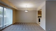 This single-floor residence offers a clean and functional layout designed for comfortable everyday living. The main living and dining area is finished with wall-to-wall carpeting in a neutral tone, creating a warm and cohesive feel throughout the space. A centrally hung pendant light fixture serves as a focal point overhead, providing both ambient illumination and a subtle design accent. The walls are painted in a soft, neutral gray, lending the space a calm and understated aesthetic. A sliding glass door along one wall invites natural light into the room and offers a connection to the outdoors. The open layout allows for flexible furniture arrangements to suit a variety of living configurations. Adjacent to the main living area, the kitchen is accessible via an open pass-through design, maintaining a sense of connectivity between spaces. The kitchen is outfitted with dark wood cabinetry, a light-toned countertop, and essential appliances including a range, making it a practical and well-equipped area for meal preparation. The overall ambiance of the space is tidy, move-in ready, and well-suited for those seeking a straightforward and functional residential environment.