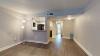 This single-floor residence offers a thoughtfully designed open layout that connects the living area to the kitchen and beyond. Rich, wood-tone laminate flooring runs consistently throughout, providing a warm and cohesive foundation across the entire space. The living area is generously proportioned, with soft gray-painted walls, white baseboards, and a textured popcorn ceiling that adds a classic character to the interior.

A defining architectural feature is the half-wall pass-through that opens into the kitchen, topped with a dark stone-style counter that functions as a breakfast bar. The kitchen is well-equipped, featuring white upper cabinetry, recessed lighting, and black appliances including a full-size refrigerator. A secondary view into the kitchen reveals a sink area with additional cabinetry and a dishwasher, offering ample workspace and storage.

A central hallway extends from the main living space toward the rear of the home, where natural light filters in through what appears to be a sliding glass door or large window, brightening the passage. The layout flows naturally from the open communal areas toward private spaces, creating a functional and comfortable living environment. Central HVAC with ceiling vents ensures year-round climate control throughout the home.