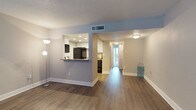This single-floor residence offers a thoughtfully designed open layout that connects the living area to the kitchen and beyond. Rich, wood-tone laminate flooring runs consistently throughout, providing a warm and cohesive foundation across the entire space. The living area is generously proportioned, with soft gray-painted walls, white baseboards, and a textured popcorn ceiling that adds a classic character to the interior.

A defining architectural feature is the half-wall pass-through that opens into the kitchen, topped with a dark stone-style counter that functions as a breakfast bar. The kitchen is well-equipped, featuring white upper cabinetry, recessed lighting, and black appliances including a full-size refrigerator. A secondary view into the kitchen reveals a sink area with additional cabinetry and a dishwasher, offering ample workspace and storage.

A central hallway extends from the main living space toward the rear of the home, where natural light filters in through what appears to be a sliding glass door or large window, brightening the passage. The layout flows naturally from the open communal areas toward private spaces, creating a functional and comfortable living environment. Central HVAC with ceiling vents ensures year-round climate control throughout the home.