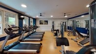 Located at 1000 Flycatcher Way in Arden, NC, this fitness center offers a comprehensive workout facility designed to support a wide range of exercise routines. The space is finished with durable cork-style flooring throughout, providing a resilient and comfortable surface underfoot. Bold navy blue accent walls contrast with crisp white trim and ceilings, giving the room a clean, energetic aesthetic. Recessed lighting and multiple ceiling fans ensure the space remains well-lit and well-ventilated during workouts.

The cardio zone is equipped with a row of treadmills, elliptical machines, and a stationary bike, all positioned to take advantage of the large windows that flood the space with natural light and offer views of the surrounding greenery. The strength training area features a multi-station cable machine, a free weight rack stocked with dumbbells, adjustable benches, a TRX suspension system, and additional resistance equipment. Full-length mirrors along one wall expand the sense of space and allow for proper form checks during exercise.

Additional amenities include a water cooler station for hydration, decorative artwork, and direct access to an outdoor area, visible through the large windows. The facility is neatly organized and thoughtfully outfitted to serve as a convenient, all-in-one fitness destination.