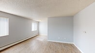 This single-floor unit offers a straightforward, functional layout highlighted by consistent wood-look flooring and soft grey walls throughout. The main living area is generously proportioned, featuring a textured popcorn ceiling, white baseboard trim, and multiple windows dressed with vertical blinds that allow natural light to filter in across the room. A wall-mounted air conditioning unit and baseboard heaters provide year-round climate control.

The galley-style kitchen is efficiently designed, featuring granite countertops, a double stainless steel sink with a modern faucet, white shaker cabinetry with gold hardware, and a full-size refrigerator. Upper cabinets provide ample storage, and the warm wood flooring carries through from the main living space.

The bathroom is clean and well-appointed, with a white vanity cabinet, a single-basin sink, gold fixtures, and a lighted mirror above. A combined tub and shower with a curtain rod is positioned adjacent to the vanity, and a window above the tub brings in natural light. The flooring matches the rest of the unit, creating a cohesive look.

Overall, the space presents a clean, move-in-ready environment with a neutral, modern aesthetic that offers a solid foundation for personalization.