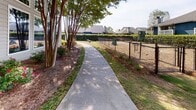 This outdoor community space is thoughtfully designed to offer a variety of amenities within a landscaped setting. Smooth concrete walkways wind throughout the grounds, connecting key areas and providing easy pedestrian access across the property. Mature trees, ornamental shrubs, and mulched garden beds line the paths, creating a well-kept and visually appealing environment.

A standout feature is the covered picnic pavilion, which provides a shaded gathering area equipped with picnic tables and a built-in grill or fireplace structure, making it well-suited for outdoor dining and social gatherings. The open lawn area adjacent to the pavilion offers additional space for outdoor activities.

A dedicated dog park is enclosed with chain-link fencing and includes pet amenities such as waste stations, offering a safe and convenient area for pets. A curved walkway bordered by crape myrtle trees and blooming rose bushes leads to this area, adding a colorful, welcoming touch.

The main building features large windows and board-and-batten style siding, framed by vibrant flowering plants. Landscaped entrance areas with decorative boulders, layered shrubs, and concrete steps complete the polished, community-oriented atmosphere of this outdoor space.