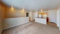 This single-floor space offers a clean, functional layout finished in warm beige tones with white trim throughout. The open living area is generously sized, featuring soft wall-to-wall carpeting and a half-wall partition that visually separates it from the kitchen while maintaining an open feel. Natural light filters through a centrally placed window, brightening the room. Ceiling-mounted light fixtures and overhead ventilation are present throughout.

The kitchen is efficiently designed with warm wood-toned upper and lower cabinetry providing ample storage. A countertop with an integrated sink, a white coil-burner electric range, and a full-size refrigerator are all included. Tile flooring runs through the kitchen, with a nearby interior door providing additional access or storage. A louvered closet door offers additional utility space within the kitchen area.

The bathroom is equipped with a single-sink vanity featuring wood cabinetry and a laminate countertop, a wall-mounted mirror with overhead lighting, a full-size soaking tub, and tile flooring consistent with the kitchen. The space is compact yet fully functional. Overall, the unit presents a straightforward, move-in-ready environment with consistent finishes and practical amenities throughout.