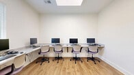 Located at 5095 Big Lake Rd in Lake Charles, Louisiana, this single-room office space offers a clean, modern, and highly functional environment designed for computer-based work. The room is thoughtfully arranged with continuous built-in, granite-topped countertop desks lining two walls, providing ample workspace for multiple users simultaneously. Each workstation is equipped with a desktop computer and monitor, paired with ergonomic rolling chairs in neutral tones accented with purple upholstery.

The space features warm wood-look luxury vinyl plank flooring that contrasts attractively against the crisp white walls and white baseboards, lending a polished and contemporary aesthetic. A window fitted with horizontal blinds allows natural light to filter into the room while offering privacy and glare control, with views of surrounding greenery visible beyond. A skylight above adds an additional source of natural illumination.

Storage is incorporated seamlessly into the design, with a gray cabinetry unit tucked beneath one section of the countertop, providing convenient closed storage. The room is accessible through a glass-paneled door, contributing to an open, professional feel. The overall ambiance is bright, orderly, and well-suited for a shared computer lab or business center environment.