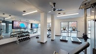 This contemporary fitness center is designed to provide a comprehensive workout experience within a clean, modern environment. The main exercise area features rubber flooring throughout and is anchored by large floor-to-ceiling windows that flood the space with natural light while offering views of the surrounding streetscape. Multiple treadmills and cardio machines, including stationary and recumbent bikes, are positioned to take full advantage of these exterior views.

Along one wall, a dedicated free weights section houses a full rack of dumbbells, complemented by large wall-mounted mirrors that enhance the sense of space and allow users to monitor their form. A multi-functional cable and strength training machine occupies a central zone, rounding out the available equipment. Two large flat-screen televisions are mounted prominently, providing entertainment during workouts.

Structural columns punctuate the open floor plan, while linear LED lighting and a ceiling fan maintain a bright, comfortable atmosphere. The space also includes a transition area near the entrance, featuring glass-paneled walls for visibility into the gym, a stainless steel waste receptacle, and patterned carpet flooring. Restroom facilities with accessible water fountains are conveniently located within the space, along with clearly marked emergency exits, ensuring both convenience and safety for all users.