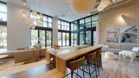Located at 3200 Paseo Village Way in San Diego, CA, this single-floor community clubhouse delivers a striking blend of modern architecture and warm, resort-inspired design. Soaring double-height ceilings define the main gathering space, which is flooded with natural light through expansive floor-to-ceiling black-framed glass walls and sliding doors that open directly to a lush outdoor pool area, bringing the Southern California landscape seamlessly indoors.

Wide-plank hardwood flooring runs throughout, anchoring the space in a natural warmth that complements the light wood paneling adorning the walls. A generously sized communal bar-height island with a two-tone wood and stone surface serves as the focal point of the central area, surrounded by woven rattan barstools. Sculptural globe pendant lights hang overhead, adding an artistic touch to the airy volume.

A full kitchenette lines one wall, featuring white cabinetry, open shelving with gold accents, and a gold-fixture sink — ideal for catered events or resident gatherings. Distinct lounge zones are thoughtfully arranged throughout, including a sectional seating area accented by curated abstract artwork on wood-paneled walls, and a cozy reading nook with a woven rattan chair framing views of the pool. The overall ambiance is sophisticated yet inviting, reflecting a carefully considered, upscale aesthetic.