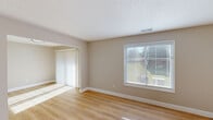 This single-floor residence offers a clean, modern aesthetic throughout, defined by warm light wood flooring, neutral warm and cool gray wall tones, and crisp white trim that create a cohesive and inviting atmosphere. The open living area is generously proportioned, bathed in natural light streaming through horizontal-slatted window blinds and vertical blind-covered sliding glass doors that invite outdoor views and a bright, airy feel. The layout flows naturally between the living and adjacent spaces, connected through wide arched passageways. The kitchen stands out as a functional centerpiece, featuring rich dark-stained cabinetry with brushed gold hardware, light quartz countertops, stainless steel appliances including a range, built-in microwave, and a sink, all illuminated by a skylight overhead that adds a distinctive architectural touch. The bedroom is well-sized with natural light from a window and offers access to a spacious walk-in closet, providing ample storage. The bathroom is finished with a full tub-shower combination, chrome fixtures, and light wood-style flooring that mirrors the rest of the home. A shiplap-accented wall in one area adds a subtle design detail. Throughout, the space presents a refreshed, move-in ready condition with consistent finishes and a neutral palette suited to a variety of personal styles.