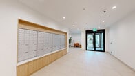 Located at 750 Robinson Street in Coquitlam, BC, this building lobby hallway serves as the primary entrance and circulation hub for residents. The space is designed with a clean, contemporary aesthetic, featuring large-format light-toned ceramic floor tiles that run throughout and bright white ceilings illuminated by recessed pot lighting, creating a consistently well-lit environment.

A prominent feature of the lobby is the expansive wall-mounted mailbox unit, elegantly framed in warm wood cabinetry. The grid of individual aluminum mailboxes offers a functional yet visually organized focal point along one wall. Adjacent to the entrance, a pair of sleek black-framed glass double doors allows natural light to filter in from the exterior, providing views of the surrounding streetscape.

The elevator area is distinguished by a warm amber-toned accent wall, providing a visual anchor alongside the stainless steel elevator doors and call panels. Potted greenery is thoughtfully placed throughout the hallway, adding organic warmth to the otherwise modern palette. A seating nook near the entrance features an upholstered armchair, offering a comfortable waiting area for residents and guests. The overall ambiance is bright, clean, and welcoming, reflecting a well-maintained, professionally designed residential building common area.