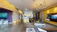 This contemporary single-floor clubhouse is designed with an open, flowing layout that seamlessly connects multiple functional zones. Polished concrete floors run throughout, lending a sleek, industrial character that contrasts beautifully with the warm, stacked-stone accent walls finished in natural limestone tones. Bold pops of color — deep purple, vibrant teal, and golden yellow — punctuate the space, adding energy and visual interest to each distinct area.

The main lounge features a dramatic multi-bulb chandelier suspended from the ceiling, casting a warm, ambient glow over comfortable seating arrangements and a wall-mounted media unit set against a rich yellow accent wall. Floor-to-ceiling glazing brings in abundant natural light and frames views of the surrounding outdoor areas. An adjacent dining zone offers a solid wood conference-style table beneath elegant pendant lights, ideal for gatherings or collaborative use.

A dedicated media lounge boasts a teal feature wall with built-in shelving and a large flat-screen TV, furnished with bold yellow accent chairs and a striped area rug. A private office nook, framed by the stacked-stone wall and trimmed in warm wood, provides a quiet workspace with a clean, minimalist desk setup. A mailroom and utility alcove with wood cabinetry and marble countertop complete the practical offerings of this well-appointed communal facility.