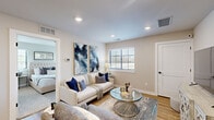 This single-floor unit offers a thoughtfully organized layout comprising two bedrooms, a living room, and a fully equipped kitchen. The living room features light hardwood flooring, recessed lighting, and multiple windows fitted with blinds that allow natural light to fill the space. Bold abstract wall art adds visual interest, while the open arrangement provides a comfortable, functional area for everyday living.

The kitchen is designed with clean white shaker-style cabinetry, black hardware accents, and light quartz countertops. Stainless steel appliances—including a range, microwave, dishwasher, and refrigerator—complement the modern aesthetic. A peninsula with seating for multiple occupants extends the counter space and creates an informal dining area. Light wood-look flooring continues from the living space, maintaining visual consistency throughout.

The primary bedroom is a well-proportioned room featuring plush carpeting, multiple windows offering ample natural light, and neutral wall tones that create a calm atmosphere. The second bedroom mirrors this restful aesthetic, with carpeted floors, a window with outdoor views, and sufficient space for standard furnishings.

Overall, the space maintains a cohesive, contemporary design defined by neutral tones, modern finishes, and efficient use of its compact footprint.