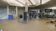 This fitness center offers a thoughtfully designed workout environment built to accommodate a variety of exercise routines. The space features a generously sized open floor plan with a durable speckled rubber flooring that provides both safety and comfort underfoot. Elevated ceilings with recessed and track lighting create a bright, energizing atmosphere throughout.

The strength training area is well-appointed with a diverse selection of resistance and weight machines, including leg press equipment, cable machines, and a variety of benches suited for multiple exercises. The machines are arranged with ample space between them, allowing for comfortable movement and use. Full-length mirrors line one side of the facility, enhancing the sense of openness while allowing members to monitor their form during workouts.

Structural columns are integrated cleanly into the layout without interrupting the flow of the space. Large windows along one wall allow natural light to filter in, contributing to the gym's open and inviting ambiance. The overall design is clean and modern, with neutral tones and a professional aesthetic that makes this fitness center a functional and motivating space for exercise.