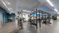 Located at 101 S Salina St in downtown Syracuse, New York, this modern fitness center offers a well-appointed, single-floor facility designed to accommodate a full spectrum of workout needs. The space features an open, expansive layout with durable rubber flooring throughout, providing both safety and comfort underfoot across all exercise zones.

The strength training area is extensively equipped with power racks, cable machines, a variety of kettlebells, medicine balls, stability balls, and TRX suspension trainers, supporting everything from functional training to traditional weightlifting. Multiple flat benches and a dedicated dumbbell section further enhance the free weights offering.

The cardio zone includes treadmills, elliptical trainers, stationary bikes, and stair-climbing machines, all positioned alongside wall-mounted flat-screen televisions for an engaging workout experience. Full-length mirrors line select walls, maximizing the sense of space and aiding users with form and technique.

Exposed ductwork and linear LED lighting run across the white ceiling, contributing to an industrial-modern aesthetic. Accent walls in muted blue and warm yellow add visual interest throughout. A glass-panel entrance connects the fitness floor to an adjacent lounge area, visible through the storefront-style doors, offering a seamless transition between the workout and relaxation spaces.