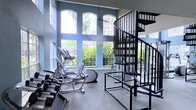 This fitness center is defined by its impressive double-height ceilings and a wall of large, arched windows that flood the space with abundant natural light while offering views of lush tropical greenery outside. The towering ceiling height creates an open, airy atmosphere that enhances the overall workout experience. The walls are finished in a cool blue-gray tone, contributing to a clean and modern aesthetic throughout the space. A striking black spiral staircase serves as a bold architectural focal point, adding a dramatic design element that contrasts with the light-filled surroundings. The flooring features a durable, speckled rubber surface well-suited for high-activity use. The gym is equipped with a selection of cardio machines, including ellipticals and step climbers, as well as a free weight rack with dumbbells, accommodating a range of fitness routines. The layout is open and functional, allowing for ease of movement between equipment. Natural light pours in throughout the day, creating an inviting environment that encourages active use. The combination of architectural character, ample equipment, and bright, green outdoor views makes this fitness center a standout amenity.