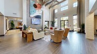 Located in Apopka, Florida, this thoughtfully designed community clubhouse offers a sophisticated and welcoming environment across a generous single-floor layout. The space is immediately distinguished by its dramatic double-height ceilings, exposed wooden beam accents, and expansive floor-to-ceiling windows that flood the interior with natural light and frame views of the surrounding outdoors.

The main lounge area serves as the heart of the space, featuring plush neutral seating arrangements, layered area rugs, and a striking mosaic tile accent wall anchored by a large mounted television and bold contemporary artwork. White-painted brick columns punctuate the open floor plan, adding architectural character while defining distinct functional zones.

A casual dining area with a long wooden table and bench seating sits adjacent to a fully equipped kitchen bar with pendant lighting, counter-height seating, and modern cabinetry — ideal for social gatherings. Wide-plank wood-look tile flooring flows seamlessly throughout, unifying the various areas with a warm, cohesive aesthetic.

Additional rooms include a dedicated game room with a billiard table and lounge seating, as well as alcove spaces offering more intimate settings. Lush indoor plants are positioned throughout, enhancing the fresh, resort-style ambiance of this well-appointed community hub.