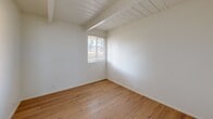 Located at 1132 Phoenix Ave in Seaside, California, this single-story, one-bedroom, one-bathroom unit offers a smartly organized layout that makes efficient use of its compact footprint. The living area features warm hardwood flooring and a distinctive wood-plank ceiling with exposed beams, lending the space a clean, understated character. A wall heater provides functional comfort throughout. The galley-style kitchen is outfitted with white cabinetry on both sides, a stainless sink with countertop workspace, and appliances including a refrigerator and range. Light tile flooring and a window overlooking the exterior keep the kitchen feeling bright and functional. A hallway connects the main living area to the bedroom, which continues the hardwood flooring and beamed ceiling aesthetic. The bedroom benefits from natural light through a window fitted with blinds and offers direct access to the bathroom. Two closets provide dedicated storage within the unit. The bathroom features tile flooring, a vanity with sink and mirror, and a window that allows natural light. The overall palette is clean and neutral throughout, with white walls creating a bright, open feel that maximizes the sense of space across every room.