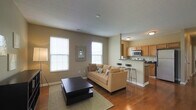 Located at 5704 New Albany Rd in West New Albany, Ohio, this single-floor residence offers a thoughtfully designed open-concept layout that seamlessly connects the living and kitchen areas. The living room is a welcoming space, featuring rich hardwood flooring, neutral taupe-toned walls, and multiple double-hung windows that fill the room with natural light. A tiered dark wood coffee table anchors the seating area, complemented by a low-profile media console and wall art, creating a balanced, contemporary aesthetic. The open-plan kitchen is positioned adjacent to the living space and is equipped with warm oak cabinetry, dark countertops, a stainless steel microwave, and a range. A raised breakfast bar with bar seating provides a convenient casual dining option and serves as a natural visual divider between the two living zones. Recessed and flush-mount ceiling lighting ensures the space remains well-lit throughout. The consistent hardwood flooring flows throughout both areas, tying the open floor plan together cohesively. Overall, the space presents a clean, functional, and comfortable living environment suited for everyday living in the New Albany community.