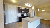 This single-story space showcases a thoughtfully designed kitchen as its centerpiece. The kitchen features rich, dark espresso cabinetry that extends to the upper walls, providing generous storage throughout. Stainless steel appliances, including a refrigerator and built-in microwave, are paired with a black range, offering a sleek, modern aesthetic contrast against the lighter surroundings.

A prominent kitchen island anchors the space, topped with a speckled granite countertop and equipped with an undermount sink and brushed nickel faucet. The island serves as both a functional workspace and a natural gathering point. Overhead, stylish cylindrical pendant lights hang at varying heights, complementing the flush rectangular ceiling fixture above the main cooking area.

Warm hardwood flooring runs throughout, adding depth and continuity to the space. The light yellow walls provide a neutral backdrop that enhances the contrast of the dark cabinetry. A hallway visible beyond the kitchen suggests additional rooms, while white panel doors and clean trim work contribute to the overall polished, well-maintained character of the space.