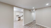 This single-floor space showcases a clean, contemporary design with a neutral color palette of soft beige and white tones carried consistently throughout. Light-toned hardwood-style flooring runs seamlessly across all areas, reinforcing a cohesive and modern aesthetic.

The open-concept layout connects a spacious main living area to the kitchen through a wide architectural opening. The living area features recessed lighting, a ceiling-mounted HVAC vent, and a closet with louvered doors for convenient storage. A separate room, accessible via its own door, includes a ceiling fan with lighting and a window fitted with blinds, providing natural light and ventilation.

The kitchen is well-appointed with white shaker-style cabinetry, granite countertops, and a gray subway tile backsplash. A full suite of stainless steel appliances includes a refrigerator, range, microwave, and dishwasher, along with a double-basin sink positioned beneath a window.

The bathroom features dark marble-style floor tile, a soaking tub with marble-surround walls, a separate glass-enclosed shower, and a granite-topped vanity, creating a polished, refined finish throughout the space.