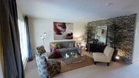 This single-floor space centers around a well-designed living room that combines contemporary and traditional elements to create an inviting, cohesive atmosphere. The room is anchored by a gray upholstered sofa dressed with decorative pillows in earthy and jewel tones, complemented by a pair of patterned accent chairs that add visual texture and character. A sleek glass-and-chrome coffee table serves as a functional centerpiece, positioned atop a woven area rug that defines the seating arrangement over neutral carpeted flooring. A standout architectural feature is the stacked stone brick accent wall, which lends warmth and a rustic-modern contrast to the otherwise light and neutral color palette of the walls and ceiling. A large framed mirror mounted on the brick wall enhances depth and reflects light throughout the space. Ambient and task lighting is thoughtfully layered, with a multi-head floor lamp and a warm table lamp contributing to a cozy, welcoming glow. Bold abstract artwork above the sofa adds an artistic focal point, while a tall potted plant near the accent wall introduces a natural, organic element to the overall design.