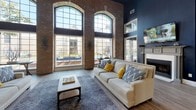 This impressive single-floor communal space showcases a bold architectural design that seamlessly blends industrial and contemporary elements. Defining the layout are prominent exposed brick columns that rise dramatically toward the soaring double-height ceilings, creating a sense of grandeur and open volume throughout. The ceiling height is accentuated further by clerestory windows positioned above the column archways, allowing additional light to filter through the upper reaches of the space.

The main lounge area features deep navy blue accent walls that provide a striking contrast to the warm tones of the brick, and is anchored by a classic white fireplace topped with a wall-mounted television. Large arched multi-pane windows line the exterior wall, offering expansive views of the outdoor pool area while bathing the interior in abundant natural light. Wood-look plank flooring runs continuously throughout, grounding the space with warmth and cohesion.

Comfortable neutral seating arrangements with coordinating blue and yellow accent pillows define the lounge area, complemented by a rustic wood coffee table and patterned area rug. A ceiling fan provides additional comfort overhead. Adjacent areas visible through open archways include a dedicated office or workstation space and what appears to be a fitness room, connected by wide, open passageways that encourage easy circulation and a connected, community-oriented atmosphere.