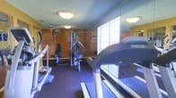 This dedicated fitness room offers a practical and functional space for exercise, outfitted with a solid selection of equipment to accommodate a variety of workout routines. The room features prominent cardio machines, including treadmills positioned near a frosted glass block window that allows natural light to filter in while maintaining privacy. A multi-station cable strength training machine anchors the center of the room, providing options for a range of resistance exercises. The flooring is finished with durable dark rubber matting, well-suited for high-traffic fitness use and equipment stability. The walls feature warm wood paneling, giving the space a classic, utilitarian character. Overhead, flush-mount ceiling light fixtures provide consistent ambient illumination throughout the room. A wall-mounted fan adds airflow and ventilation support. The textured ceiling adds a traditional finishing touch. Decorative wall accents lend a welcoming feel to an otherwise purpose-driven environment. Overall, the fitness room is thoughtfully arranged to maximize usable floor space while keeping all equipment accessible, making it a convenient and complete facility for daily exercise needs.