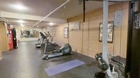 This single-level fitness room offers a practical and well-equipped space for residents or users seeking a convenient workout facility. The room features exposed concrete block walls painted in a neutral beige tone, giving it a utilitarian character typical of a building amenity space. The flooring consists of dark rubber gym matting, which provides durability and traction during exercise. A row of cardio machines, including ellipticals and a treadmill, lines one side of the room, creating a dedicated aerobic workout zone. A yoga mat is laid out on the floor, designating an area for stretching or floor exercises. On the opposite end, weight training equipment, including a cable machine and free weights, rounds out the strength training offerings. A wall-mounted air conditioning unit provides climate control, while recessed and overhead lighting keeps the space adequately illuminated. Framed informational or motivational posters are mounted along the walls, adding a modest decorative touch. A mirrored wall near the entrance enhances the sense of depth and allows users to monitor their form during workouts. Overall, the space is straightforward, functional, and geared toward everyday fitness needs.