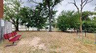 Located in Odenton, Maryland, this community dog park offers a dedicated outdoor space designed for pets and their owners to enjoy together. The park is enclosed by a chain-link fence with a gated entry, ensuring a secure environment for dogs to roam freely. The ground cover consists of open grassy and bare-earth terrain, providing ample room for dogs to run and play. Several large, mature trees are distributed throughout the park, offering natural shade and a pleasant canopy overhead. A set of colorful agility equipment is positioned centrally within the park, giving active dogs an outlet for structured play and exercise. Near the entrance gate, a waste disposal station is conveniently placed, complete with a green waste bin and an informational signage post to help keep the area clean and well-maintained. The park is bordered by a brick building on one side, while views of the surrounding residential neighborhood are visible through the fence line. The overall atmosphere is open, casual, and community-oriented, making it a practical and welcoming destination for local dog owners in the Odenton area.