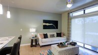 This well-appointed single-floor residence offers a thoughtfully designed living space that balances modern aesthetics with everyday comfort. The living room serves as the heart of the home, showcasing soft sage-green walls that create a calm, welcoming atmosphere. Light hardwood flooring runs throughout, adding warmth and a sense of continuity to the space. A large sliding glass door with patterned drape panels draws in abundant natural light while providing access to the outdoors, enhancing the open and airy feel of the room. The living area is anchored by a crisp white sofa adorned with an eclectic mix of decorative pillows in earthy and navy tones, complemented by a textured mosaic-style coffee table that adds visual interest. A bold abstract canvas in dark tones serves as a striking focal point on the main wall, adding a touch of artistic character. A round side table paired with a warm table lamp contributes to the layered lighting scheme, while a ceiling-mounted light fixture provides overhead illumination. The colorful area rug introduces playful accents of teal, coral, and orange, tying the design elements together. Overall, the space exudes a contemporary yet inviting ambiance, ideal for both relaxing and entertaining.