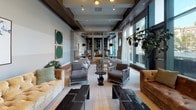 This thoughtfully designed single-floor space functions as an expansive communal lounge, seamlessly integrating multiple seating areas within one open, flowing layout. The interior is anchored by warm hardwood flooring that runs throughout, complemented by a neutral palette of gray, taupe, and white, accented with rich earth tones through furnishings.

Floor-to-ceiling windows line the perimeter, flooding the space with abundant natural light and offering views of the surrounding streetscape. Sheer white curtains soften the light while maintaining an airy, open feel. Dark wood beam detailing frames the elevated ceilings, adding architectural depth and contrast.

A cozy lounge nook features a tufted golden velvet sofa paired with a dark marble coffee table and a built-in wall unit housing a large flat-screen television, open shelving filled with sculptural décor, and curated accessories. A striking butterfly artwork and a tall floor lamp add warmth to this corner.

The café-style dining area offers banquette seating with mosaic tile detailing, café tables, and pendant lighting. Additional seating clusters throughout the space include upholstered armchairs in gray and deep green velvet, arranged to encourage both intimate conversation and open socializing, creating a refined yet welcoming environment.