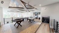 This expansive single-floor amenity space spans over 2,500 square feet, thoughtfully designed to accommodate both social gatherings and recreational activities. The layout is anchored by two distinct living areas, each carrying its own character while sharing a cohesive modern farmhouse aesthetic throughout.

The primary lounge features soaring ceilings with decorative wood beam inlays, shiplap accent walls, and striking chandelier lighting. Geometric herringbone and parquet-patterned flooring transitions seamlessly between zones, adding visual depth throughout. Large windows invite abundant natural light and neighborhood views, framed by bold plaid drapery. A statement tiered chandelier and barrel-style arcade game contribute to the room's eclectic charm.

The game room is a standout feature, outfitted with a foosball table, shuffleboard, and a dedicated golf simulator bay complete with a projection screen and turf mat. An arched passageway connects the lounge areas, while hallways lead to elevator access and additional amenities including a bar-height counter and kitchenette.

The covered outdoor terrace extends the living space further, featuring slatted wood deck tiles, ceiling fans, multiple dining and lounge seating arrangements, and open views—making it an ideal setting for year-round gatherings.