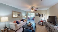 This single-floor residence is thoughtfully designed with a cohesive coastal aesthetic throughout. The living room serves as the heart of the home, featuring soft sage-green walls, plush carpeting, and a ceiling fan overhead. A large sliding glass door floods the space with natural light and opens directly onto a private balcony, offering pleasant outdoor views. A decorative mirror and framed wall art enhance the room's character.

The galley-style kitchen is efficiently laid out with warm honey-toned wood cabinetry, dark granite-style countertops, a double-basin sink, and stainless steel appliances including a refrigerator and electric range. Rich hardwood flooring runs underfoot, adding warmth to the space.

Adjacent to the kitchen, the dining area accommodates a round wooden table beneath a pendant light fixture, creating an intimate setting for meals. Neutral walls and carpeted flooring carry through into the generously sized bedroom, which features a ceiling fan, natural window light, and direct access to a private en-suite bathroom. The bathroom includes wood-toned cabinetry, a tub-shower combination, and hardwood flooring. A dedicated in-unit laundry area with stacked washer and dryer completes the residence.