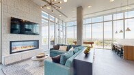 This high-rise amenity floor is a masterclass in contemporary design, seamlessly blending open, light-filled gathering spaces with functional work and entertainment areas. Floor-to-ceiling glass curtain walls wrap the main lounge, flooding the space with natural light and framing expansive panoramic views of the city skyline and lush treetop canopy below. Soaring ceilings amplify the sense of openness, while a sculptural sputnik chandelier adds a bold mid-century modern accent overhead.

The central lounge area is anchored by a dramatic floor-to-ceiling textured stone fireplace with a linear gas insert and wall-mounted display screen, surrounded by plush teal sectional seating and a marble-topped coffee table. Large-format porcelain tile flooring grounds the space with a clean, refined aesthetic. Direct access to an outdoor terrace extends the living area, where additional lounge seating offers alfresco enjoyment with unobstructed views.

The kitchen features a substantial quartz-topped island with a fluted tile base, warm wood cabinetry, marble-look backsplash, stainless steel appliances, and a striking slatted wood ceiling detail that adds architectural warmth. Adjacent conference rooms enclosed in sleek glass partitions with dark metal framing provide dedicated spaces for meetings, equipped with wall-mounted displays, beverage refrigerators, and contemporary furnishings — ideal for professional gatherings within this elevated setting.