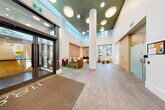 Located in Vancouver, BC, this residential building lobby offers a well-designed, contemporary entrance experience across a generously sized single floor. The space is immediately distinguished by its dramatic double-height ceilings, which create an open, airy atmosphere and allow natural light to pour in through an upper clerestory window band. The ceiling is finished in a bold deep green, contrasting elegantly with the crisp white walls and smooth cylindrical structural columns that anchor the space. Underfoot, large-format stone-look tile flooring extends throughout the main circulation areas, transitioning to green textured area rugs that define the furnished lounge zones. The central gathering area features a warm wood bar-height table paired with modern white molded chairs, ideal for residents to meet and collaborate. A separate lounge nook is furnished with a distinctive channeled tan leather sofa, a round white coffee table, and warm wood wall paneling, offering a more relaxed setting. Circular flush-mounted ceiling fixtures provide even, ambient lighting throughout. Additional functional features include parcel lockers, a resident bulletin board, staircase access, and secured entry doors with glazed sidelights. The overall ambiance is refined, welcoming, and thoughtfully appointed.