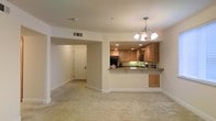 This single-floor residence offers a thoughtfully designed open-concept layout that seamlessly connects the living and kitchen areas. The living space is finished with soft, neutral-toned carpeting that adds warmth and comfort underfoot, while crisp white walls and clean baseboards create a bright, airy atmosphere throughout.

The kitchen is positioned at the heart of the home, separated from the living area by a peninsula counter topped with granite-style stone that provides both additional prep space and casual seating. Medium-toned wood cabinetry lines the upper and lower walls of the kitchen, offering generous storage. Recessed lighting illuminates the workspace efficiently, complemented by stainless steel appliances including a microwave and refrigerator. A green-tinted tile backsplash adds a subtle accent of color to the cooking area.

A decorative chandelier with a classic multi-arm design serves as a focal point overhead in the dining and kitchen zone, adding an elegant touch to the space. A hallway visible from the main living area leads to additional rooms, providing a clear and intuitive flow throughout the floor plan. HVAC ventilation is integrated into the ceiling, ensuring climate comfort. Overall, the space presents a clean, functional, and welcoming environment.