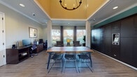 Located at 70 Mowry Ave in Fremont, California, this single-story community clubhouse is designed to serve as a versatile gathering and productivity hub. The space is anchored by a dramatic tray ceiling finished in warm golden yellow and adorned with a statement chandelier, creating an impressive sense of vertical volume throughout the main room.

At the center of the space sits a large solid-wood communal table with modern wire-frame chairs, suitable for meetings, collaborative work, or social gatherings. To one side, a dedicated workstation area features computer desks and ergonomic chairs, providing a functional business center for residents. Opposite the workstations, a floor-to-ceiling bank of smart package lockers offers a modern mail and delivery solution.

A comfortable lounge area is positioned near the large windows, furnished with tufted swivel accent chairs and a decorative side table, bathed in abundant natural light from multiple oversized windows framed by elegant drape panels with blinds. The views to the surrounding greenery and an outdoor pool area enhance the connection between interior and exterior spaces. Glass-panel doors provide direct access to the pool deck, further integrating the indoor and outdoor environments.

Throughout, warm hardwood-style flooring, crown molding, structural columns, and neutral wall tones contribute to a cohesive, polished aesthetic that balances comfort with functionality.