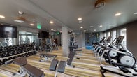 Located at 2025 Welton St in Denver's historic Five Points neighborhood, this fitness center offers a comprehensive workout facility spread across a single level. The main gym floor is generously proportioned, featuring striped carpeting and recessed lighting complemented by ceiling fans that maintain a comfortable environment during workouts. Large windows line the perimeter, flooding the space with natural light and providing views of the surrounding urban landscape.

The main floor is equipped with a wide array of strength and cardio equipment, including multiple treadmills, ellipticals, cable machines, free weights, and adjustable benches. A standout feature is the colorful rock climbing wall, which adds a dynamic and unique training option. Mirrored walls expand the sense of space and allow for form monitoring during exercise.

A separate studio room features dark hardwood-style flooring, full-length mirrors, spin bikes, step platforms, and slam boxes, making it ideal for group or solo high-intensity training. Ceiling fans keep the space ventilated. An additional transitional area with dark tile flooring includes a wall-mounted television and vibration platforms. Storage shelving, a small refrigeration unit, and towel storage are conveniently available throughout the facility.