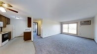 This single-floor residence offers a clean, functional layout designed to maximize comfort and usability. The open-concept living area is finished with soft, neutral-toned carpeting that adds warmth underfoot, while bright white walls and a smooth ceiling create a fresh, airy atmosphere throughout. A large sliding glass door floods the living space with abundant natural light and provides direct access to an outdoor balcony, offering views of the surrounding landscape. The kitchen is positioned adjacent to the living area and is defined by rich dark wood cabinetry that provides a warm contrast to the lighter tones of the space. It features a stainless steel sink and extends toward a hallway that leads to additional rooms. The vinyl flooring in the kitchen area transitions seamlessly into the carpeted living space, creating a natural separation of zones. A corridor visible from the main living area connects to further rooms, suggesting a thoughtfully arranged floor plan. The overall ambiance of the unit is light, open, and neutral — well-suited for comfortable everyday living. The combination of natural light, practical finishes, and an open layout makes this a welcoming and versatile residential space.