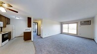 This single-floor residence offers a clean, functional layout designed to maximize comfort and usability. The open-concept living area is finished with soft, neutral-toned carpeting that adds warmth underfoot, while bright white walls and a smooth ceiling create a fresh, airy atmosphere throughout. A large sliding glass door floods the living space with abundant natural light and provides direct access to an outdoor balcony, offering views of the surrounding landscape. The kitchen is positioned adjacent to the living area and is defined by rich dark wood cabinetry that provides a warm contrast to the lighter tones of the space. It features a stainless steel sink and extends toward a hallway that leads to additional rooms. The vinyl flooring in the kitchen area transitions seamlessly into the carpeted living space, creating a natural separation of zones. A corridor visible from the main living area connects to further rooms, suggesting a thoughtfully arranged floor plan. The overall ambiance of the unit is light, open, and neutral — well-suited for comfortable everyday living. The combination of natural light, practical finishes, and an open layout makes this a welcoming and versatile residential space.