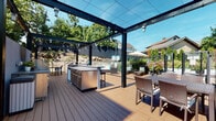Located at 120 N Pearl St in Port Chester, NY, this thoughtfully designed outdoor deck offers a sophisticated al fresco living experience. The space is anchored by a striking steel-framed pergola with cable wire detailing and string lights overhead, creating an inviting atmosphere for daytime and evening gatherings alike. Underfoot, warm-toned composite decking runs throughout, lending a clean and durable foundation to the entire area. At the heart of the deck sits a fully equipped outdoor kitchen, complete with a stainless steel island, built-in appliances, and bar seating — ideal for entertaining. Multiple dining and lounge zones are thoughtfully arranged across the deck, furnished with wicker seating sets and complemented by lush plantings and a patio umbrella for shade. The perimeter is defined by modern wire mesh railings with a sleek dark metal frame, offering unobstructed views of the surrounding mature trees and landscaping while maintaining a sense of enclosure and safety. Steps lead to an adjacent lower deck area, expanding the overall outdoor footprint. The contemporary facade of the neighboring building, clad in warm terracotta panels, adds an architectural backdrop to the setting. This outdoor space seamlessly blends functionality and design for year-round outdoor living.