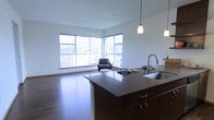This single-floor space showcases an open-concept layout that seamlessly connects the kitchen and living areas. The kitchen is anchored by a substantial dark-finished island with a stainless steel sink, rich cabinetry with brushed hardware, and a pendant light overhead, offering both functionality and a strong visual focal point.

The living area benefits greatly from a pair of large corner windows that span nearly floor to ceiling, flooding the space with natural light and providing open views of the surrounding urban landscape. The expansive glazing creates an airy, connected feel between the interior and the city beyond.

Warm-toned hardwood flooring runs throughout, adding continuity and a sense of cohesion to the entire open floor plan. The clean white walls keep the space feeling bright and neutral, complementing the contrast of the dark kitchen finishes.

Overall, the space delivers a modern, urban aesthetic with a practical, flowing layout suited to everyday living and entertaining, characterized by natural light, quality finishes, and a well-considered open design.