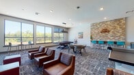 This game room is a dynamic, multi-functional recreational space designed to foster social interaction and entertainment. The room is unified by a patterned carpet that adds visual texture throughout. Large windows line the walls, flooding the space with natural light and offering open views of the surrounding greenery.

The billiards area serves as a central focal point, featuring a full-size pool table with a sleek pendant light overhead. A cue rack stands nearby, and the space is complemented by a dining-height table with modern chairs for casual seating. Adjacent to the pool table, a stacked-stone accent wall adds a warm, rustic architectural element, anchoring a high-top bar counter with teal-colored stools.

A dedicated TV lounge occupies one corner of the room, where a deep burgundy accent wall provides a bold backdrop for two wall-mounted flat-screen televisions. Comfortable brown leather lounge chairs and ottomans are arranged to create an inviting viewing area, with additional high-top seating positioned near the windows.

A ping pong table is positioned in an open area of the room, providing ample space for play. The room also features recessed lighting throughout and direct access to adjacent areas via interior doors, making it a versatile and engaging space for recreation and socializing.