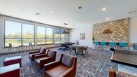 This game room is a dynamic, multi-functional recreational space designed to foster social interaction and entertainment. The room is unified by a patterned carpet that adds visual texture throughout. Large windows line the walls, flooding the space with natural light and offering open views of the surrounding greenery.

The billiards area serves as a central focal point, featuring a full-size pool table with a sleek pendant light overhead. A cue rack stands nearby, and the space is complemented by a dining-height table with modern chairs for casual seating. Adjacent to the pool table, a stacked-stone accent wall adds a warm, rustic architectural element, anchoring a high-top bar counter with teal-colored stools.

A dedicated TV lounge occupies one corner of the room, where a deep burgundy accent wall provides a bold backdrop for two wall-mounted flat-screen televisions. Comfortable brown leather lounge chairs and ottomans are arranged to create an inviting viewing area, with additional high-top seating positioned near the windows.

A ping pong table is positioned in an open area of the room, providing ample space for play. The room also features recessed lighting throughout and direct access to adjacent areas via interior doors, making it a versatile and engaging space for recreation and socializing.