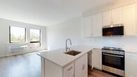This well-designed single-floor unit presents a clean, contemporary aesthetic with a bright and airy atmosphere throughout. The open-concept layout seamlessly connects the kitchen and living area, creating a cohesive and functional living space.

The kitchen is a standout feature, outfitted with crisp white shaker-style cabinetry that extends to the ceiling, offering generous storage. A central island with a light quartz countertop includes an undermount stainless steel sink with a modern pull-down faucet, providing both prep space and a natural divider between the kitchen and living area. Stainless steel appliances — including a range, over-the-range microwave, dishwasher, and full-size refrigerator — complete the kitchen. A white subway tile backsplash adds a clean, classic touch.

Light hardwood-style flooring flows consistently throughout the main living spaces, contributing to the unit's warm yet modern feel. Large windows in the living area flood the space with natural light and offer views of the surrounding neighborhood. The bedroom is a calm, neutral retreat with the same flooring and clean white walls.

The bathroom is finished with wood-look tile flooring, white cabinetry, and minimalist fixtures, maintaining the consistent modern design language found throughout the unit. Overall, the space offers a fresh, move-in-ready environment with thoughtful finishes and an efficient, open layout.