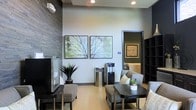 This single-floor space is thoughtfully designed to create a professional yet comfortable environment. The waiting area serves as the focal point, combining modern aesthetics with a calming atmosphere ideal for a professional or medical setting. The room features light hardwood-style flooring that contrasts elegantly with the dark slate-textured accent wall, adding depth and visual interest to the space. Recessed ceiling lighting provides even, warm illumination throughout. Seating consists of contemporary upholstered chairs in neutral gray tones, arranged around dark-finished side tables, offering guests a comfortable place to wait. A water cooler station is conveniently positioned along the back wall, adding a practical touch. The décor is anchored by two large nature-inspired artworks depicting soft, muted tree branches, contributing to the serene and tranquil ambiance. Potted plants, including a striking dark-leafed agave, introduce natural elements that enhance the overall freshness of the space. A darkly framed doorway leads to an adjacent room, suggesting additional functional areas beyond the waiting zone. The overall design balances professionalism with warmth, making it a welcoming environment for visitors.
