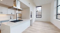 Located at 165 Richards St in West Haven, CT, this thoughtfully designed one-bedroom, one-bathroom unit offers a clean, modern aesthetic with high-quality finishes throughout. Light hardwood flooring flows seamlessly across the entire space, complementing the crisp white walls and creating a bright, cohesive feel.

The open-concept kitchen is a standout feature, showcasing white shaker cabinetry with sleek matte black hardware, quartz countertops, a central island with a gas cooktop, a stainless steel refrigerator, dishwasher, and a statement stainless steel range hood overhead. The kitchen opens directly into the living area, which is anchored by a dramatic floor-to-ceiling black-framed window that floods the space with natural light and offers views of the surrounding neighborhood.

The hallway connects the main living areas to the private quarters, lined with louvered closet doors that provide practical storage. The bedroom is a serene retreat featuring its own large black-framed window with neighborhood views and continued hardwood flooring. The bathroom is finished with large-format neutral tile on the walls and floors, a soaking tub with shower, and recessed lighting for a spa-like ambiance. An in-unit laundry area adds everyday convenience to this well-appointed home.