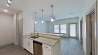 This well-designed single-floor residence offers a clean, contemporary aesthetic throughout, highlighted by light wood-look flooring, crisp white walls, and abundant natural light. The open-concept kitchen is a standout feature, boasting white shaker-style cabinetry, granite countertops, a spacious peninsula with an undermount sink, and a full suite of stainless steel appliances including a refrigerator, range, microwave, and dishwasher. Elegant pendant lighting hangs above the peninsula, complementing the track lighting along the kitchen corridor. The kitchen flows seamlessly into a bright living area equipped with a ceiling fan and multiple windows, along with a glass door that invites natural light and outdoor views. The bedrooms are comfortably sized, each featuring ceiling fans, windows with blinds, and the same warm wood-look flooring found throughout the home. The primary bathroom offers a dual-sink vanity with granite countertops, a well-lit mirror, and access to a walk-in closet. A second bathroom provides a full tub and shower combo with clean, simple finishes. The layout, as seen in the dollhouse view, is thoughtfully arranged for efficient flow and functionality across all living spaces.